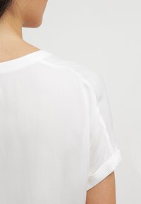 Weiße Bluse aus weichem, leichtem Stoff, mit kurz geschnittenen Ärmeln, die an den Bündchen hochgerollt sind, und einem lässigen Schnitt. Details umfassen einen runden Halsausschnitt.