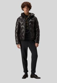 Mustaparkainen toppatakki, jossa on siksak-kuvio, vetoketjusuljenta ja huppu. Päälle puettu tumma neule ja harmaat housut, yhdistetty tummiin kenkiin.