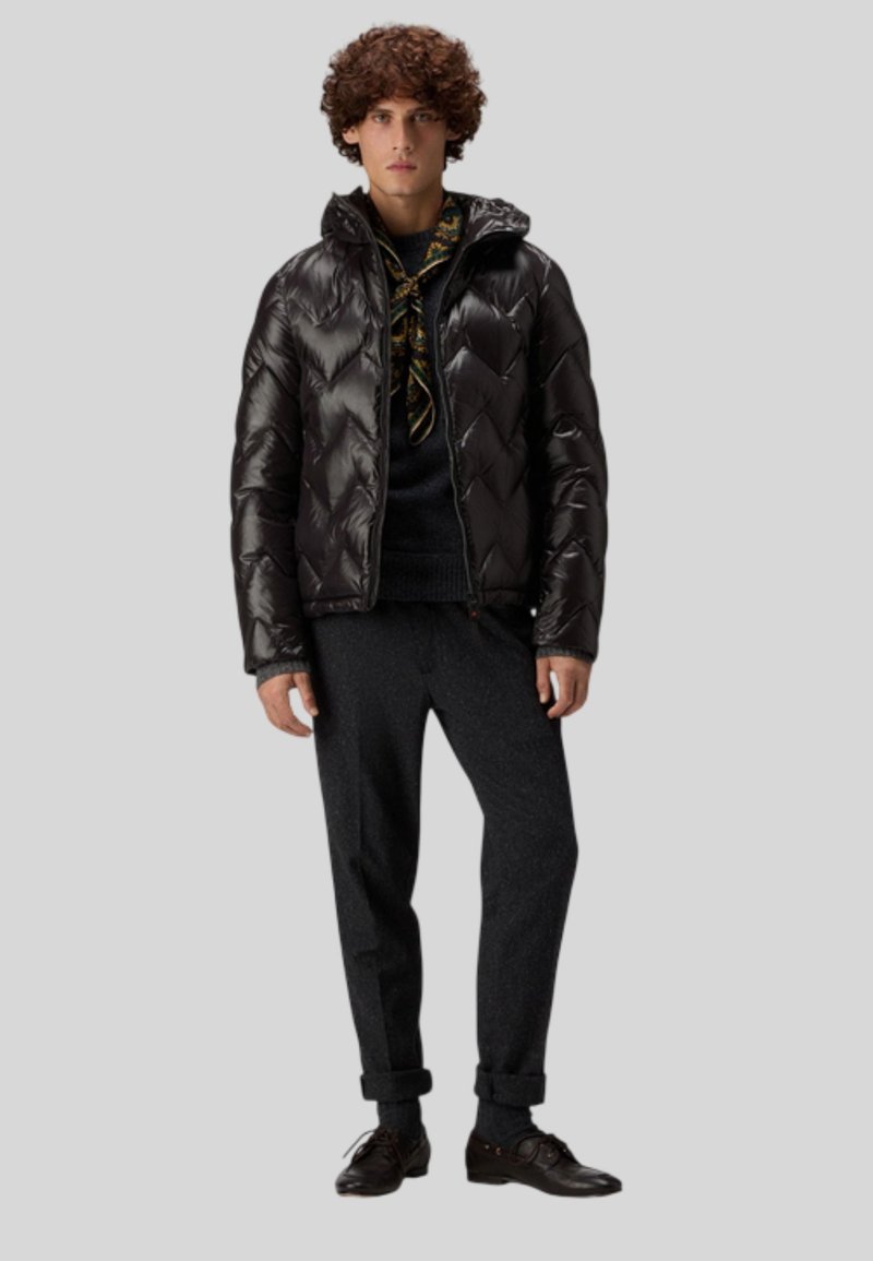 Mustaparkainen toppatakki, jossa on siksak-kuvio, vetoketjusuljenta ja huppu. Päälle puettu tumma neule ja harmaat housut, yhdistetty tummiin kenkiin.