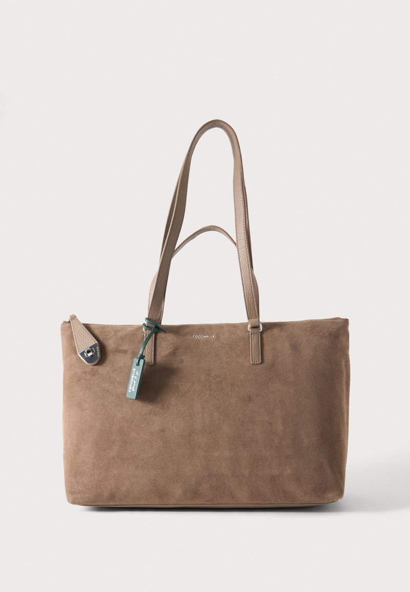 Sac cabas en daim marron avec deux longues poignées, petit logo sur le devant et une étiquette verte suspendue sur un fond clair uni.