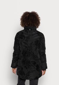 Svart puffy jacka med hög krage, med ett strukturerat blommönster och kviltad design. Långa ärmar och en böjd fåll bak.