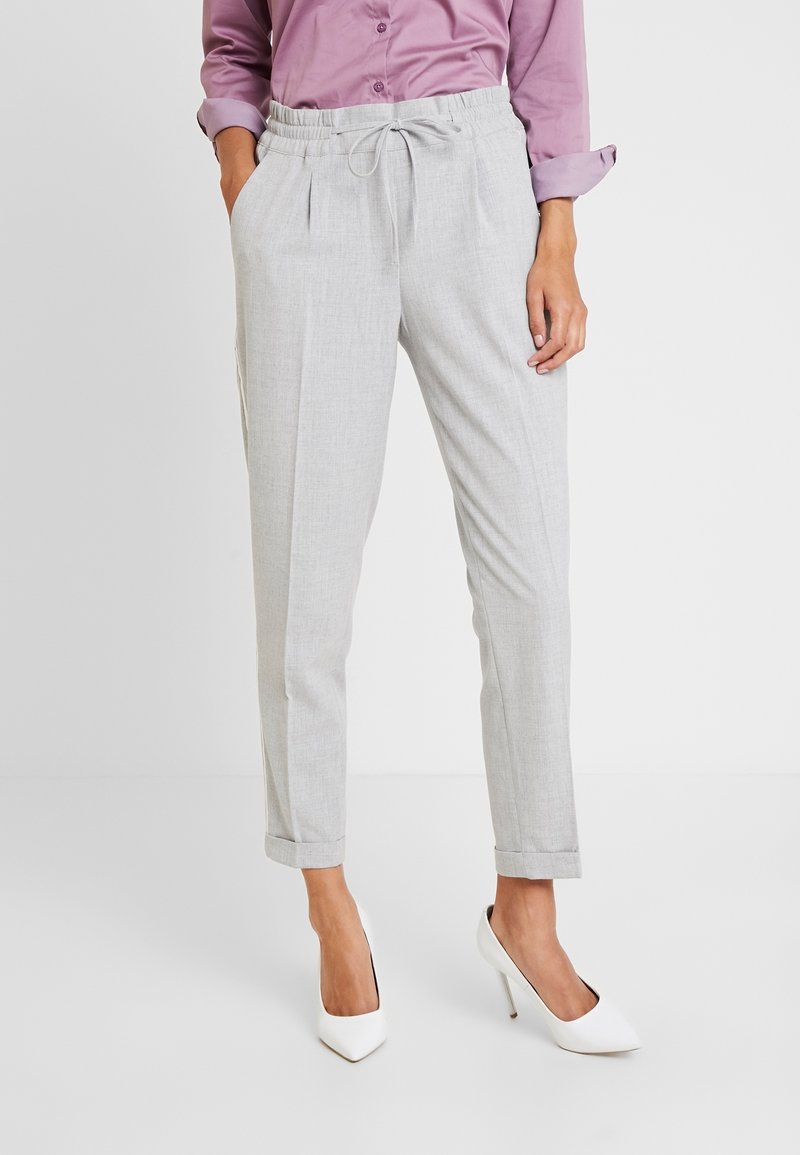 Femme portant un pantalon gris clair fuselé avec une taille à cordon, associé à une chemise mauve à manches longues et des chaussures blanches à talons hauts.