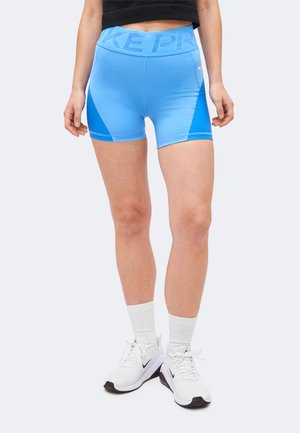 Vrouw die lichtblauwe sportshorts met meshpanelen, witte Nike-sneakers en witte enkelsokken draagt, staand tegen een lichte achtergrond.