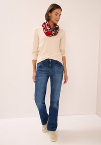 Un haut beige à manches longues associé à un jean en denim bleu, comportant cinq poches. Une écharpe à motifs multicolores est portée autour du cou.