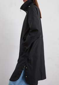 Imperméable noir avec col haut, zip avant intégral, poches latérales et boutons-pression. Présente une texture lisse et une silhouette fluide.