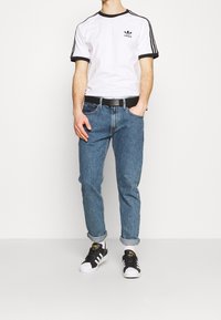 Bílý tričko s krátkým rukávem, černými akcenty a logem, modré džíny, ohrnuté manžety a černé tenisky s bílou špičkou. Součástí je černý opasek.