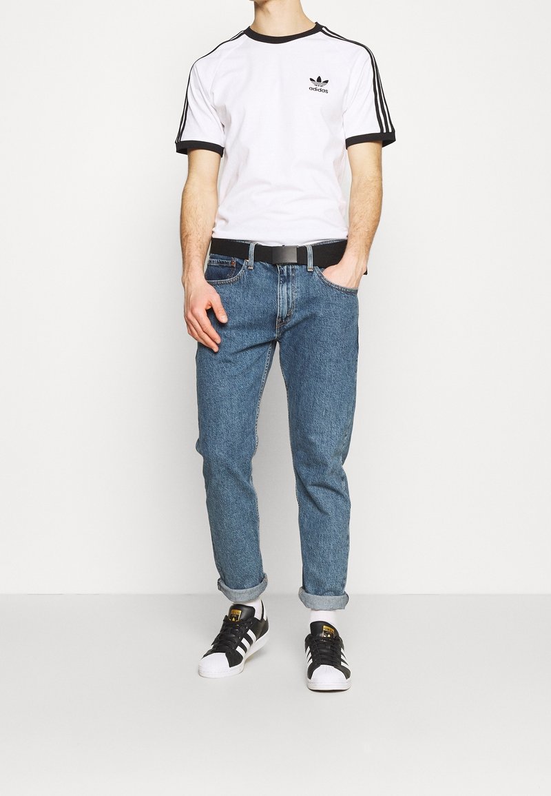 Bílý tričko s krátkým rukávem, černými akcenty a logem, modré džíny, ohrnuté manžety a černé tenisky s bílou špičkou. Součástí je černý opasek.