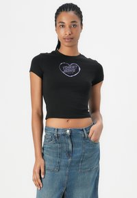 Svart avkortet t-skjorte med korte ermer, med en lilla og hvit "TOMMY JEANS"-logo. Kombinert med høy midje denimskjørt.