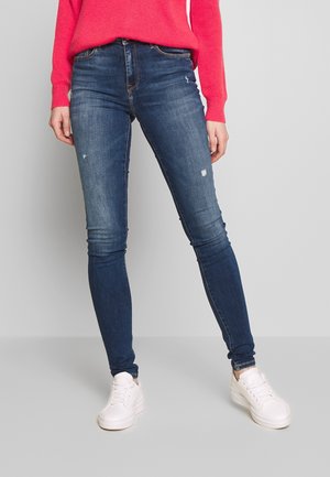 Jean skinny bleu foncé avec un léger délavage et des détails usés, associés à des baskets blanches et un pull rose vif.
