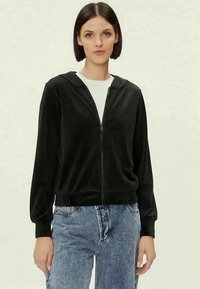 Eine Frau trägt einen schwarzen Reißverschluss-Hoodie über einem weißen Shirt und blauen Jeans und steht vor einem schlichten hellen Hintergrund.