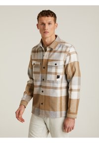 Camisa de franela a cuadros en beige, marrón y blanco; cuenta con dos bolsillos en el pecho con cierres de botón y un parche de logo en el bolsillo izquierdo.
