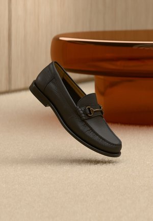 Mocassino in pelle nera con dettaglio a morsetto in metallo, posizionato sopra un tappeto beige, con un tavolo rotondo marrone sullo sfondo.