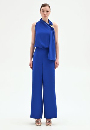 Donna che indossa una tuta senza maniche blu royal con dettaglio a sciarpa drappeggiata e spilla dorata, abbinata a sandali metallici aperti, in piedi su sfondo bianco.