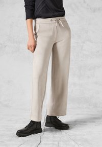 Beige vide benbukser lavet af glat stof, med elastisk talje og sidelommer, parret med sorte snørebåndsstøvler.