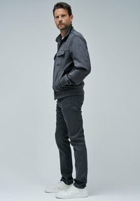 Chaqueta bomber de ante gris con un bolsillo frontal, combinada con pantalones gris ajustados y zapatillas blancas, sobre un fondo neutro.