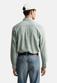 Grünes, strukturiertes Langarmhemd, Rückansicht, kombiniert mit blauen Jeans und einer Kappe. Auffällige Details: geknöpfte Manschetten und ein gerader Schnitt.