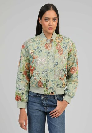 Femme aux longs cheveux foncés porte une veste bomber vert clair avec des motifs floraux colorés et un jean bleu, debout devant un fond gris.