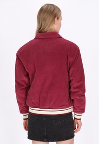 Giacca in corduroy bordeaux con texture a coste, dotata di colletto, polsini e orlo a righe, dalla vestibilità rilassata, indossata sopra una gonna di jeans nera.