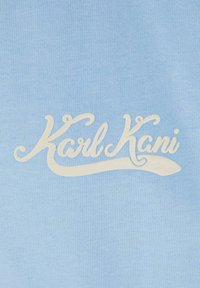 Šviesiai mėlynas medvilnės audinys su baltu siuvinėtu "Karl Kani" logotipu, parašytu kursyvu, pasižymintis glotna tekstūra ir subtiliu blizgesiu.