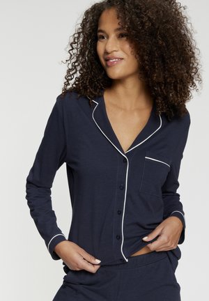 Žena s kučeravými vlasmi, ktorá má na sebe tmavomodré pyžamo na zapínanie s bielym lemovaním, jemne upravuje lem košele a jemne sa usmieva, stojac pred jednoduchým pozadím.