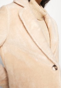 Abrigo de piel sintética marrón claro con cuello de muesca, cierre de un solo botón y textura suave. Llevado sobre un suéter de cuello alto de canalé en color beige claro.