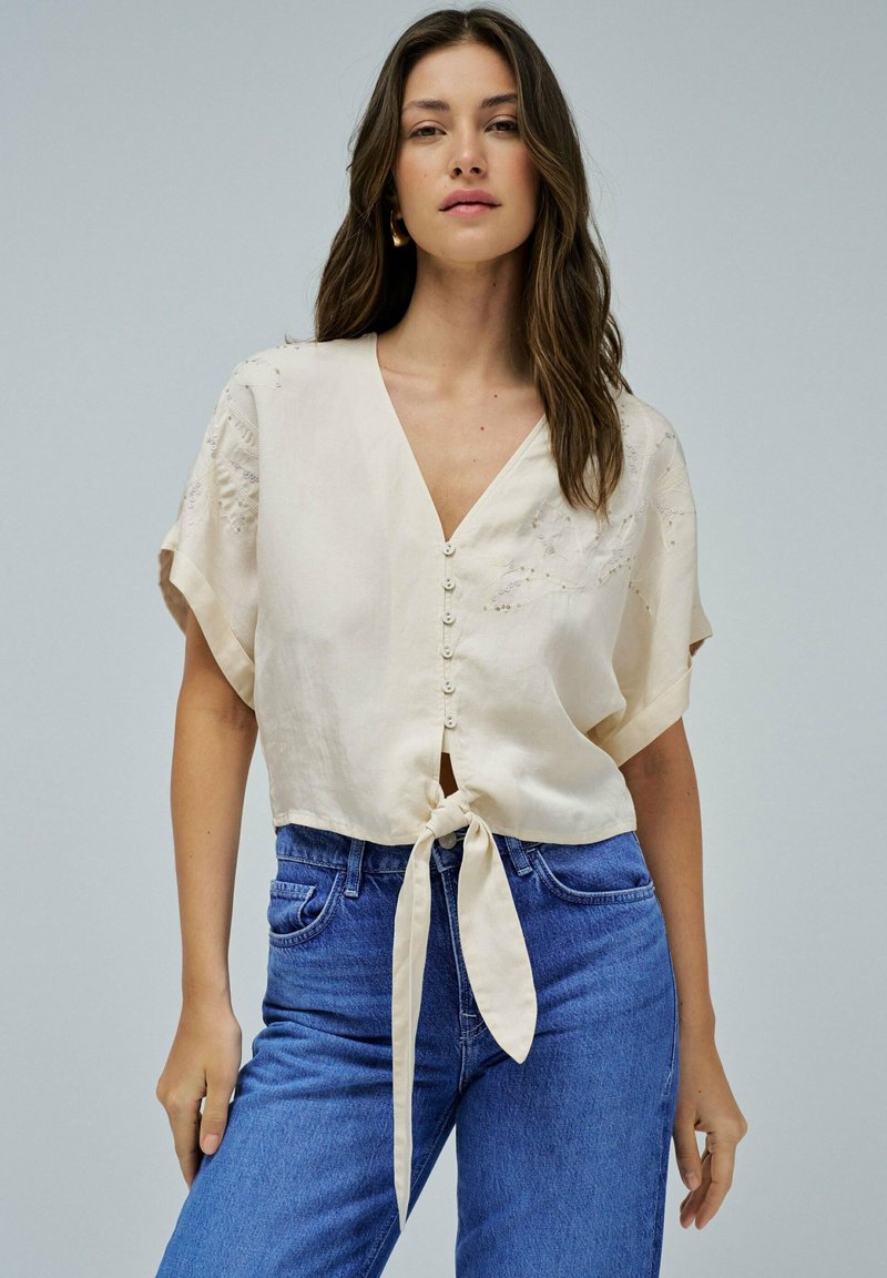 Blusa accorciata color crema con scollo a V, maniche corte e un nodo frontale. Presenta bottoni al centro e delicati dettagli ricamati.