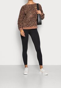 Persona con sudadera marrón con estampado de leopardo, leggings negros, zapatillas blancas y llevando un bolso negro de hombro, de pie contra un fondo blanco.