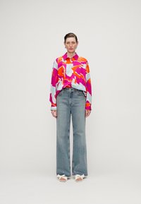 Colorful button-up shirt with bold pink, orange, and light blue patterns, paired with high-waisted light denim jeans and white sandals.
