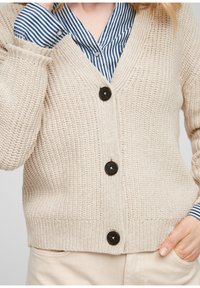 Krémový pletený kardigan s výstřihem do V, se třemi tmavými knoflíky, žebrovaným lemem a manžetami. Pod ním je vidět modrá a bílá pruhovaná košile.