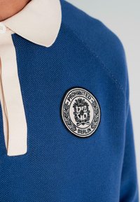 Blue knit polo shirt with cream collar featuring a round black and white "Prohibited Berlin" patch on the chest.