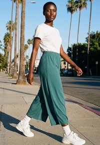 Young woman walking on sunny sidewalk lined with tall palm trees, wearing white shirt, teal wide-leg pants, white socks, and white sneakers.