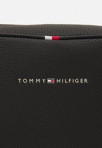 Sort tekstureret lædertaske med en silder logo plade, der læser "TOMMY HILFIGER" og en detalje med et rødt, hvidt og blåt accentstribe.