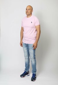 T-shirt rosa chiaro a maniche corte con un piccolo logo scuro, abbinata a jeans in denim blu chiaro e sneakers sportive blu navy con dettagli rossi e bianchi.