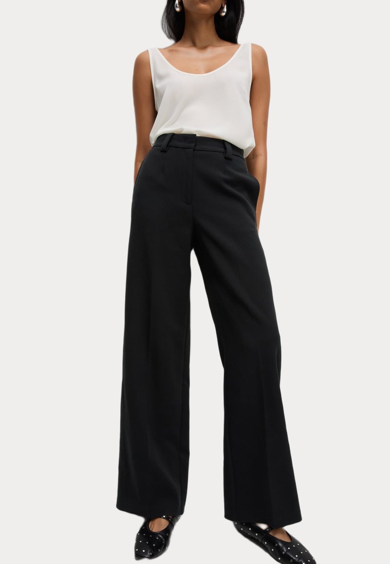 Femme portant un haut blanc sans manches, un pantalon noir taille haute à jambes larges, et des ballerines noires cloutées, debout devant un fond uni.