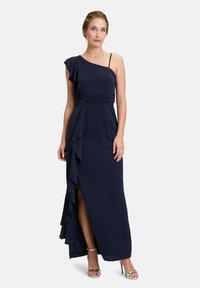 Mörkblå, enaxlad aftonklänning med volangdetalj, rynkad midja och hög slits, kompletterad med bandiga silverskor.