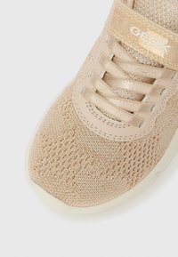 Zbliżenie różowego dzianinowego trampeca z brokatowymi detalami, satynowymi sznurówkami i błyszczącym paskiem z napisem "GEOX" na białym tle.