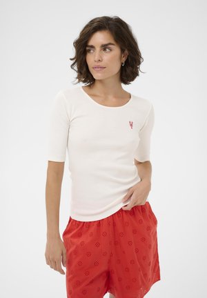Femme aux cheveux courts bruns portant un haut blanc à manches trois-quarts avec un petit emblème de homard rouge et un short rouge à motifs, posant devant un fond uni.