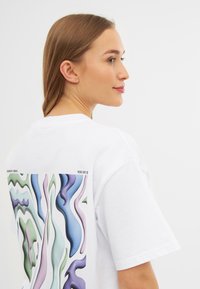 Camiseta blanca con un diseño abstracto colorido en tonos de azul, verde y púrpura en la parte posterior. Mangas cortas y corte relajado.