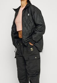 Black quilted bomber jacket with a zip front, ribbed cuffs, and a small white logo. Paired with black cargo pants featuring pocket details.