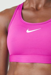 Tuvumā redzams cilvēks ar spilgti rozā Nike sporta krūšturi, redzama augšējā ķermeņa daļa un logo uz krūtīm.