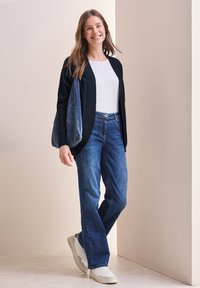 Un cardigan noir avec des bords côtelés, un t-shirt blanc, un jean bleu taille haute et des baskets blanches. Un sac en denim pend à l'épaule.