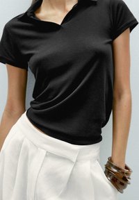 Black polo shirt with a collar, featuring a relaxed fit. Worn with white pleated trousers and brown bangle bracelets, against a light backdrop.
