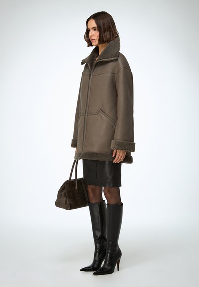 Abrigo de piel vuelta marrón con cuello alto, cierre de cremallera y bolsillos delanteros; combinado con una falda de cuero negra, botas negras hasta la rodilla y un bolso marrón.