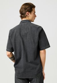 Camicia in denim grigio scuro a maniche corte con colletto classico, cuciture visibili e design della spalla posteriore. Il tessuto presenta una texture sottile.