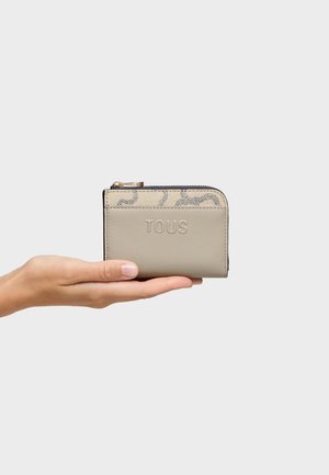 Mano sosteniendo una pequeña cartera beige con cremallera y el logo en relieve "TOUS" sobre un fondo neutro.