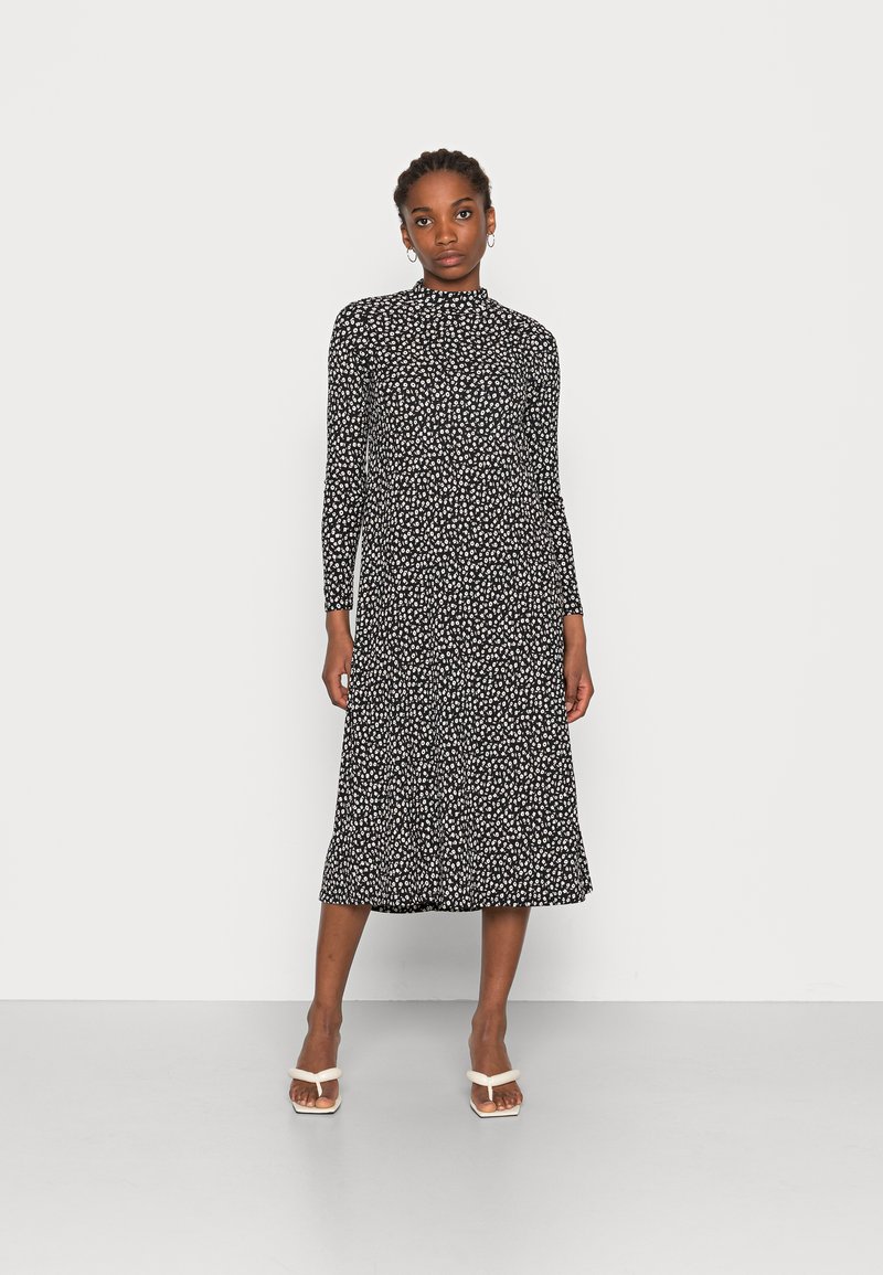 Vestido preto de mangas longas com estampa floral branca, decote alto, comprimento midi fluido e tecido suave. Combinado com sandálias brancas de tiras.