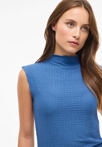 Blå ärmlös topp med en hög, vågig krage. Strukturerad tyg med ett subtilt mönster, figurnära design som framhäver kroppen.