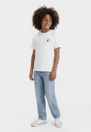 Kind met krullend haar, gekleed in een wit T-shirt met een klein rood logo, lichtblauwe spijkerbroek en witte sneakers, staand en glimlachend.