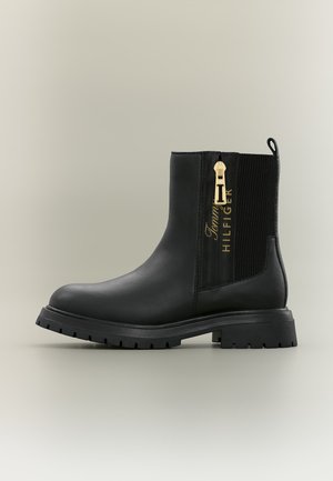 Schwarzer Knöchelstiefel mit klobiger Sohle, goldenem Reißverschluss an der Seite, elastischem Panel und "Tommy Hilfiger" Logo in goldenen Lettern.