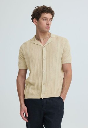 Ung mann iført en beige kortermet, knappeopp strikkeskjorte og svarte bukser, ser til venstre med en hånd i lomma.